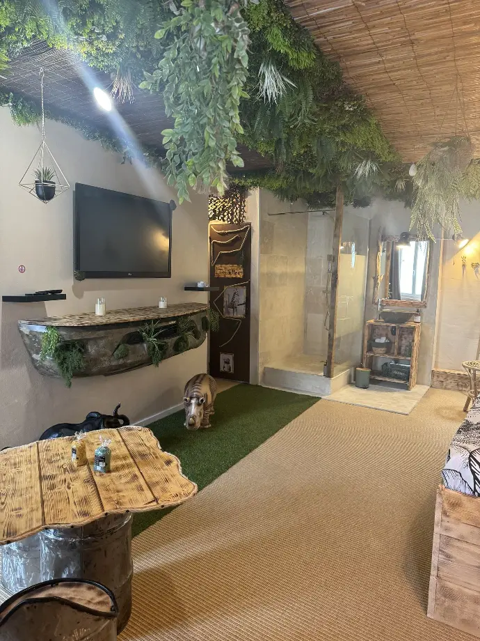 Suite La Jungle de Sissy avec plafond végétalisé, télévision murale, salle de bain ouverte, et décoration jungle avec statuette d'hippopotame et table en bois naturel Locations de Sissy, Saintes-Maries-de-la-Mer