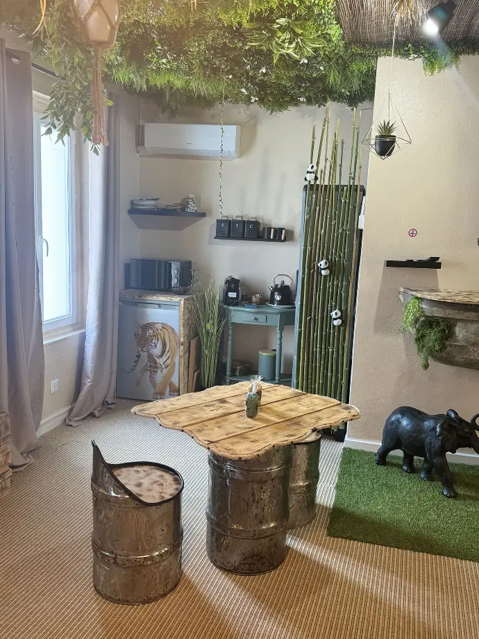 Espace de détente dans la suite La Jungle de Sissy avec table en bois, tabourets en métal, décorations exotiques, réfrigérateur, climatisation, et plafond végétalisé Locations de Sissy, Saintes-Maries-de-la-Mer