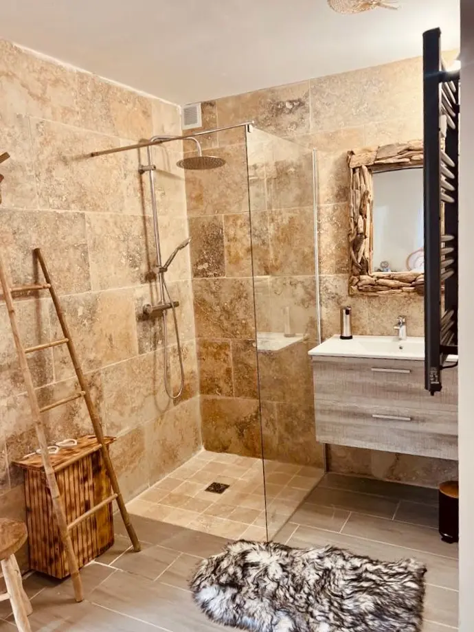 Douche à l'italienne dans salle de bain en pierre naturelle aux Locations de Sissy avec miroir en bois flotté et tapis en fausse fourrure.